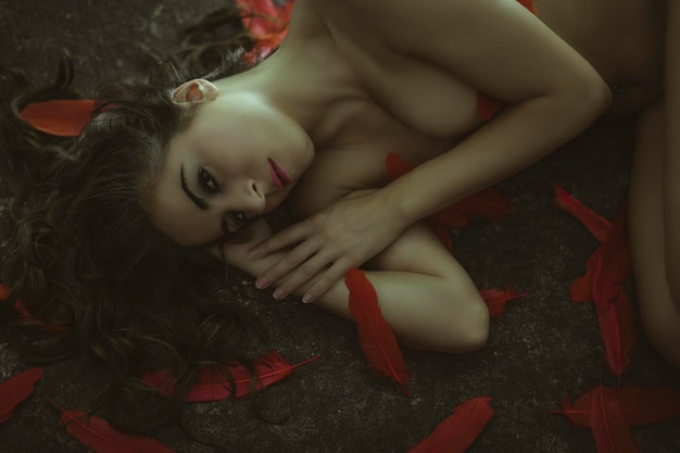 Sensuality, Sensual nude brunette woman with long black hair, lying on the ground surrounded by red bird feathers