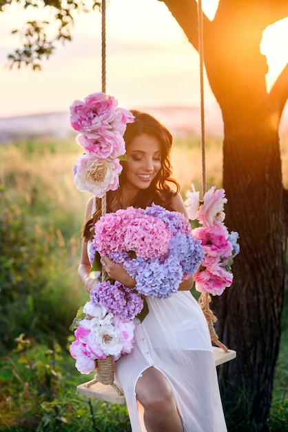 Sensueel mooi meisje met perfecte glimlach in een witte jurk zittend op een houten schommel met een boeket van tedere gekleurde bloemen.