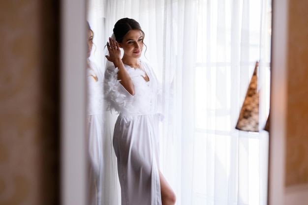 Sensuele bruid jonge vrouw met perfecte bruiloft kapsel en make-up poseren in de buurt van groot raam Aantrekkelijke blanke bruid meisje gekleed in witte peignoir trouwdag partij voorbereiding