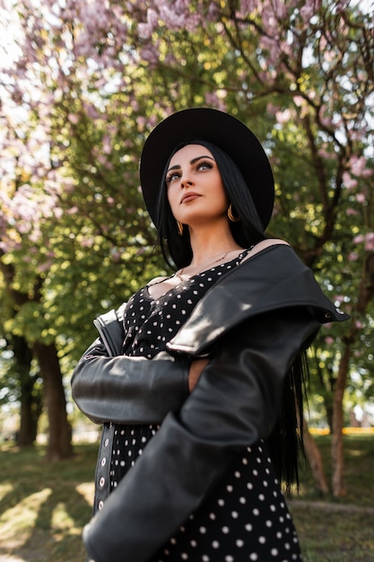 Sensuele jonge vrouw in zwarte vintage jurk in mooie stijlvolle hoed in trendy leren jas staat in het park op de achtergrond van lila bomen. mooie sexy meisje in elegante slijtage in zonnige dag in de natuur. kunnen.