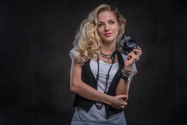 Sensuele krullend blond meisje fotograaf gekleed in een wit t-shirt en vest draagt veel accessoires en polshorloge, poseren met een camera in een studio. Geïsoleerd op donkere gestructureerde achtergrond.