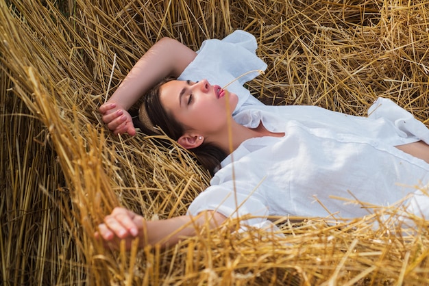 Sensuele vrouw die in het hooi ligt Meisje in wit overhemd en ligt op een hooiberg Vrouwelijk portret in het veld op het platteland