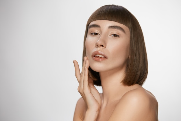 Foto sensuele vrouw met perfecte huid aanraken van haar gezicht, brunette met perfecte rechte korte haarstijl en blote schouders met glanzende lippen op witte achtergrond