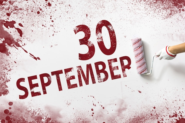 9月30日。月の30日、カレンダーの日付。手は赤いペンキでローラーを保持し、白い背景にカレンダーの日付を書き込みます。秋の月、年の日の概念。