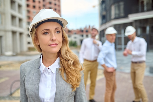 Безмятежная привлекательная женщина-инспектор в каске стоит на строительной площадке с рабочими-мужчинами на заднем плане