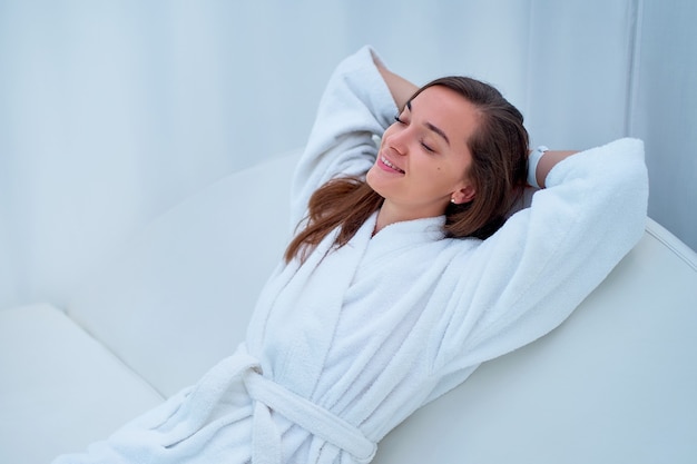 Serene calm woman wearing white bathrobe with closed eyes and hands behind head enjoying relaxing time and feeling good at a wellness spa resort.