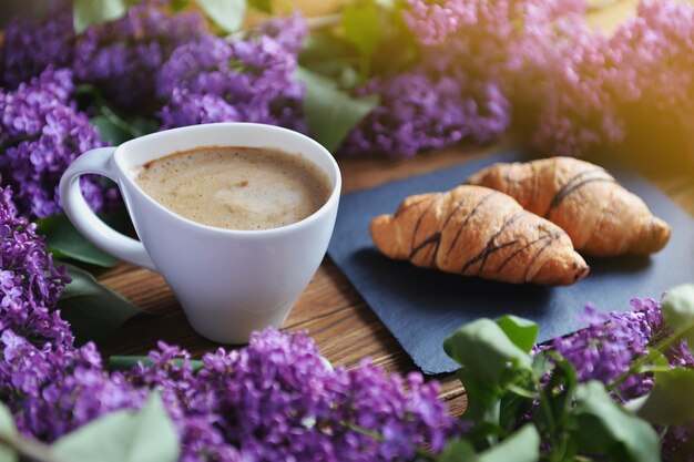 Serene croissants and a cappuccino cup are on the table. A bouquet of lilac on a wooden table.