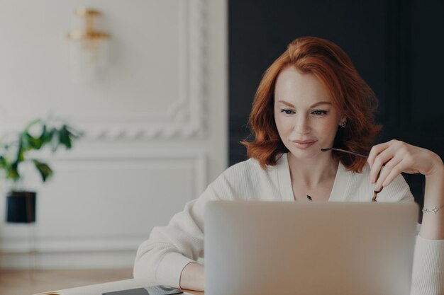 Спокойная рыжая с очками, работающая на ноутбуке, одетая в шикарный белый свитер в изысканном домашнем офисе