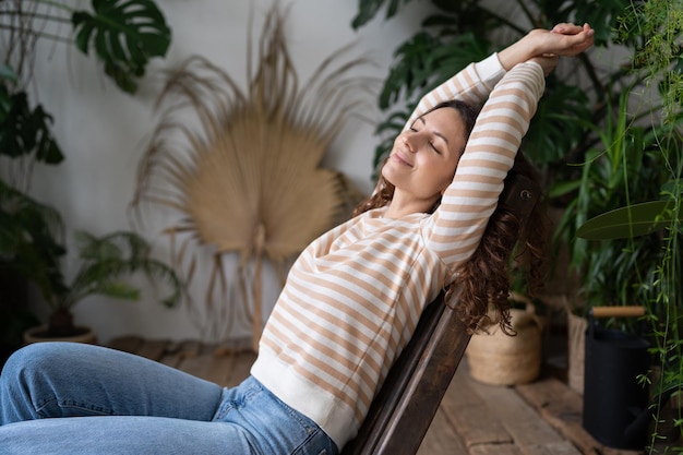 Serene relaxed young woman with closed eyes stretching arms relaxing in urban jungle interior