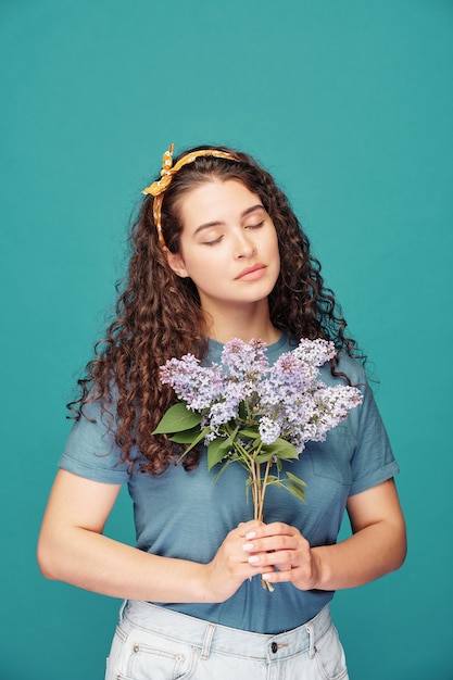 Serene romantic girl with curly hair keeping eyes closed and holding purple flowers on blue