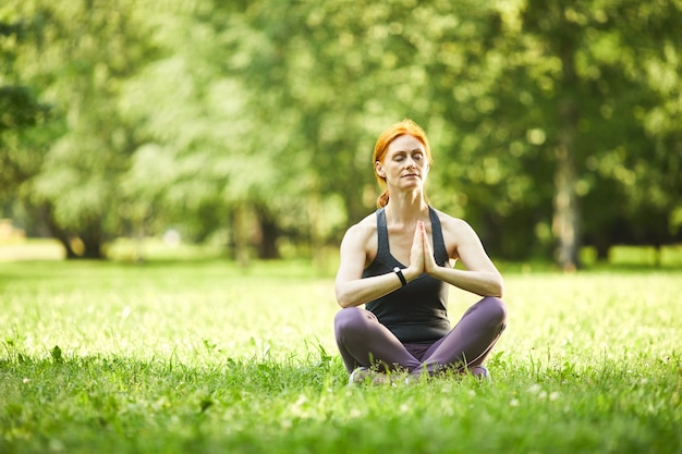 Serene roodharige rijpe vrouw zittend op het gras en hand in hand in namaste tijdens het mediteren in een leeg park