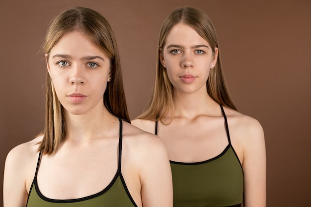 Photo serene twins in pistachio underwear tanktops looking at camera while standing against brown background in isolation