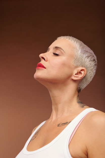 Serene woman with short blond hair, closed eyes, tattoos on neck and chest and red lipstick on lips expressing pleasure in isolation