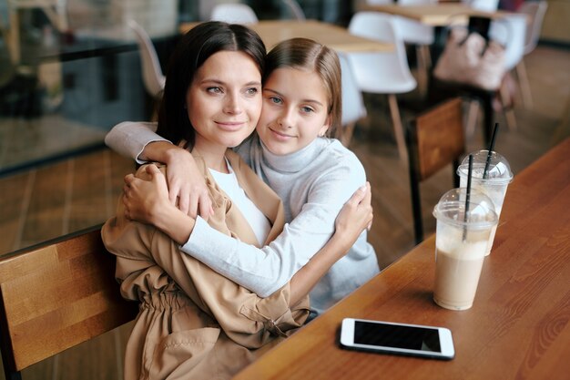 Serena giovane donna e sua figlia seduti in un abbraccio mentre ci si rilassa nella caffetteria e sorseggiando un cocktail di latte