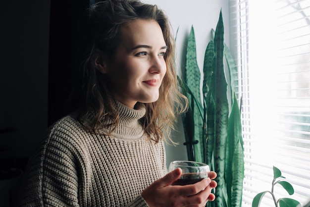 A serene young woman savors a hot cup of tea while admiring the stunning cityscape from a window