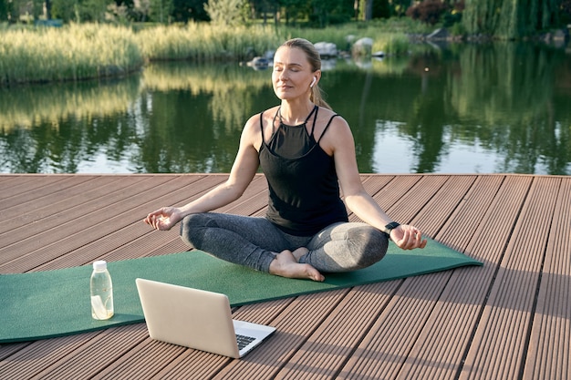 Serenity sporty relaxed woman meditating sitting in lotus pose while watching tutorial on a laptop