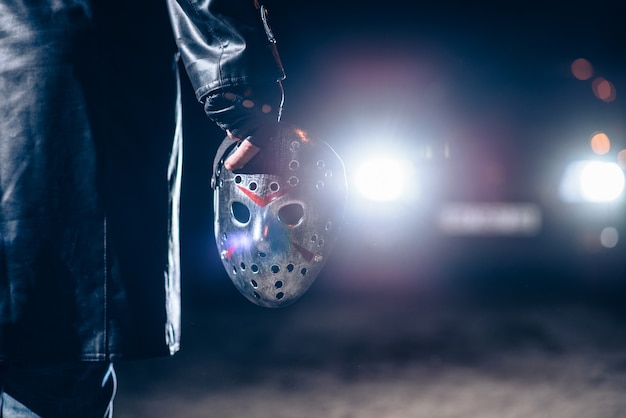 Serial maniac hand with bloody hockey mask closeup, car light at the night. Horror, bloody murderer