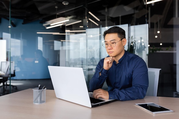 Serieus denkende aziatische zakenman die werkt in een moderne kantoor volwassen man in shirt en bril