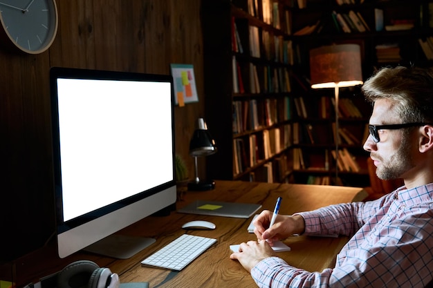 Serieus gefocuste jonge zakenman die een desktopcomputer gebruikt en naar een mock-up wit wit schermmonitor kijkt voor reclame, laat werkt, online leert op een website, webinar kijkt op zijn kantoor thuis.