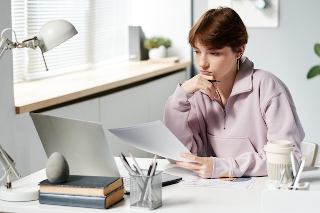 Serieus Kaukasisch meisje in hoodie zit aan het bureau en kijkt naar het laptopscherm terwijl ze het budget analyseert