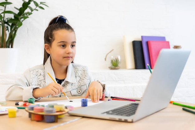 Serieus klein meisje met koptelefoon handgeschreven studie online met behulp van laptop thuis, schattig gelukkig klein kind neemt internetwebles of klas op pc.