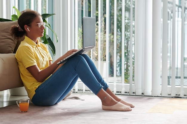 Serieus nieuwsgierig gemengd ras meisje in bril zittend op de vloer en met behulp van moderne laptop tijdens het kijken naar educatieve video