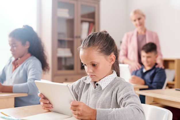 Serieus schattig meisje en haar klasgenoten met digitale tablets die bij bureaus zitten en individueel werken met online tekst terwijl de leraar hen raadpleegt