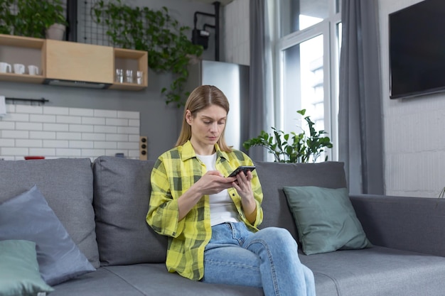 Serieuze en gefocuste vrouw die thuis op de bank zit en het nieuws van de telefoon leest