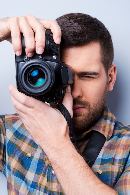 Serieuze fotograaf. Portret van een zelfverzekerde jonge man in shirt die camera voor zijn gezicht houdt terwijl hij tegen een grijze achtergrond staat