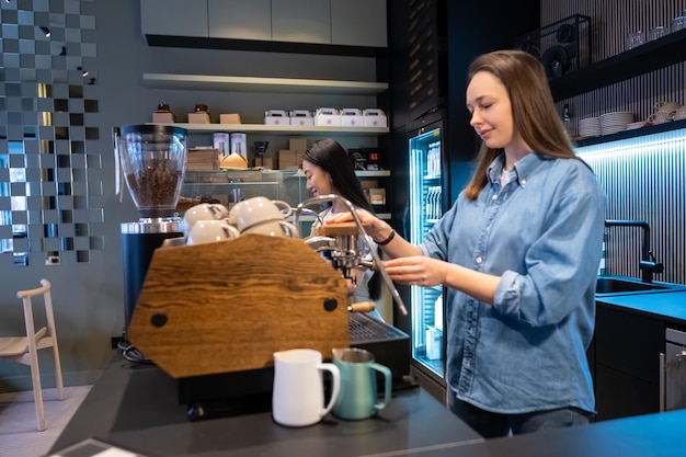 Serieuze geconcentreerde blanke barista die de koffieautomaat bedient naast haar tevreden Aziatische vrouwelijke collega.