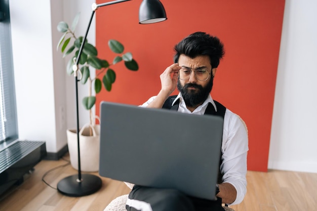 Serieuze Indiase zakenman in formele kleding die aan het typen is op een laptop terwijl hij een zachte poef zit in een licht coworking-kantoor bij het raam