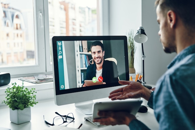 Serieuze jonge man die met een collega praat via een videogesprek terwijl hij op kantoor zit