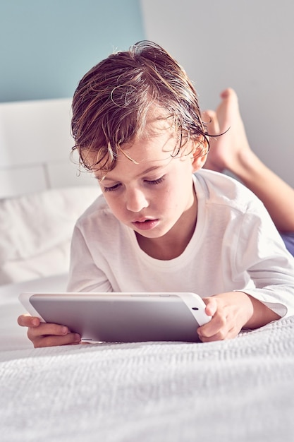 Serieuze jongen met nat haar na de douche ligt op bed en kijkt naar cartoons op tablet in de slaapkamer