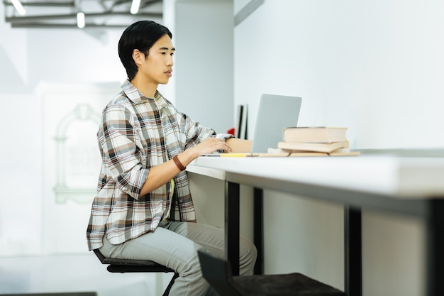 Serieuze man. Rustige geconcentreerde Aziatische persoon zit aan het bureau in een moderne coworking-ruimte en werkt op zijn laptop