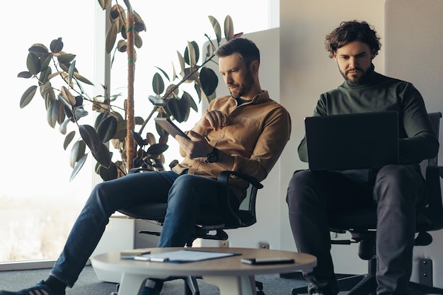 Serieuze mannelijke collega's die samenwerken met behulp van laptopcomputer en digitale tablet op het hedendaagse bedrijfskantoor