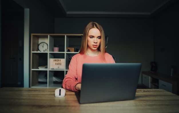 Foto serieuze mooie vrouw in een roze trui zit thuis aan een tafel en werkt online op een laptop