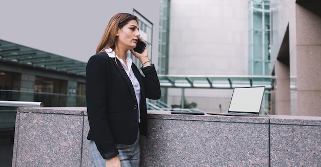 Serieuze vrouwelijke manager die in de buurt van een stenen hek staat en wegkijkt tijdens het beantwoorden van een telefoontje