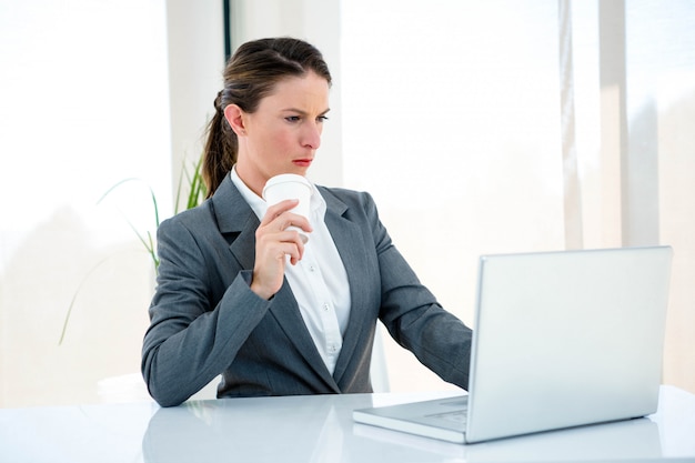 serieuze zakenvrouw, achter haar bureau, te typen op haar laptop en koffie te drinken