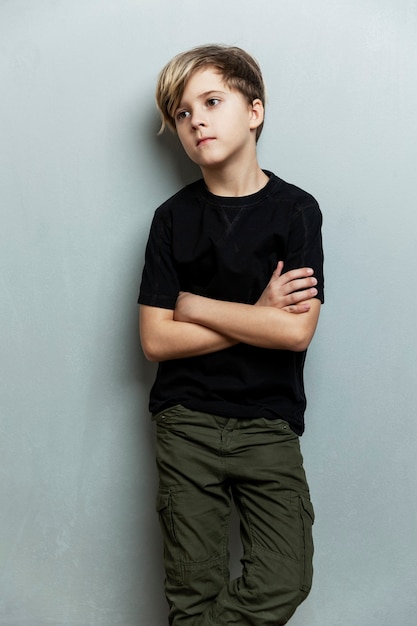 黒のTシャツを着た真面目な9歳の少年が壁に立っています。腕は胸に交差しています。灰色の背景。垂直。