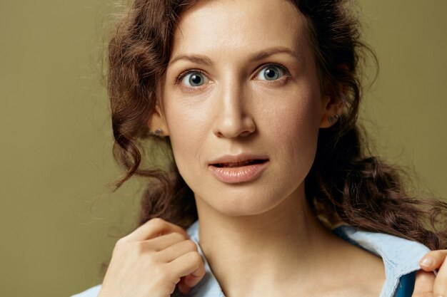 Serious adorable curly beautiful female in jeans casual shirt\
touches collar looks at camera posing isolated on olive green\
background being yourself people lifestyle emotions concept copy\
space