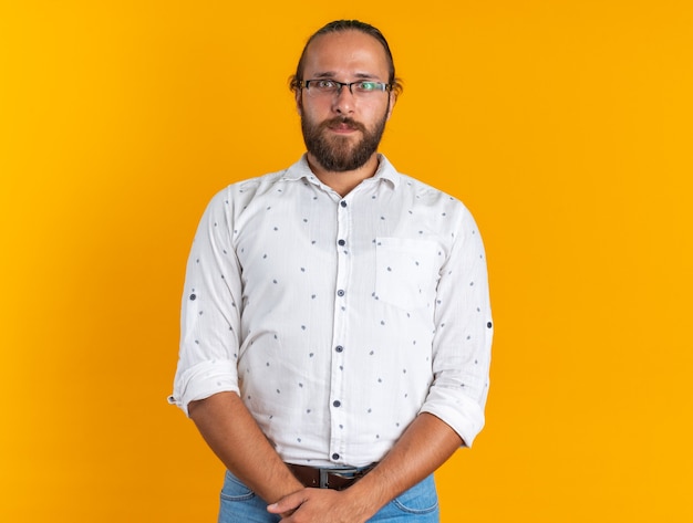 Serious adult handsome man wearing glasses keeping hands together looking at camera isolated on orange wall