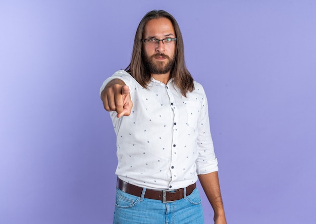 Serious adult handsome man wearing glasses looking and pointing at camera isolated on purple wall with copy space