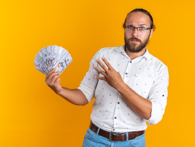 Serious adult handsome man wearing glasses showing money looking at camera showing four with hand isolated on orange wall