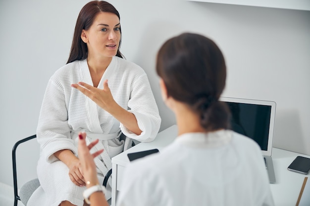 Serious adult woman in a bathrobe complaining about her skin problems to a qualified dermatologist