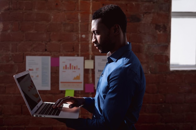 Serious african american businessman standing in office holding and using laptop. working in business at a modern office.