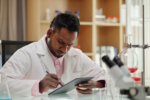 Serious african american clinician or researcher using tablet by workplace in laboratory while searc