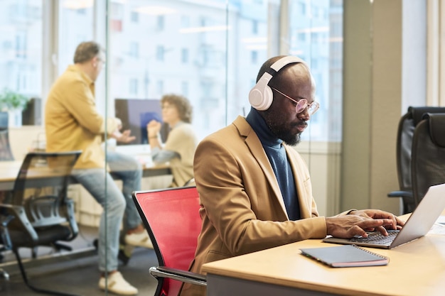 Serious African American employee with headphones typing by workplace