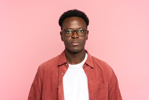 Serious African American man in glasses wear red shirt, looking at camera standing isolated on pink