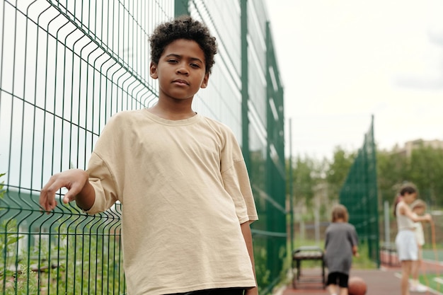 Serious african american schoolboy in beige tshirt standing by fence