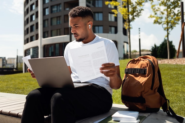 Serious african american student guy using laptop computer outdoors and looking at papers sitting in